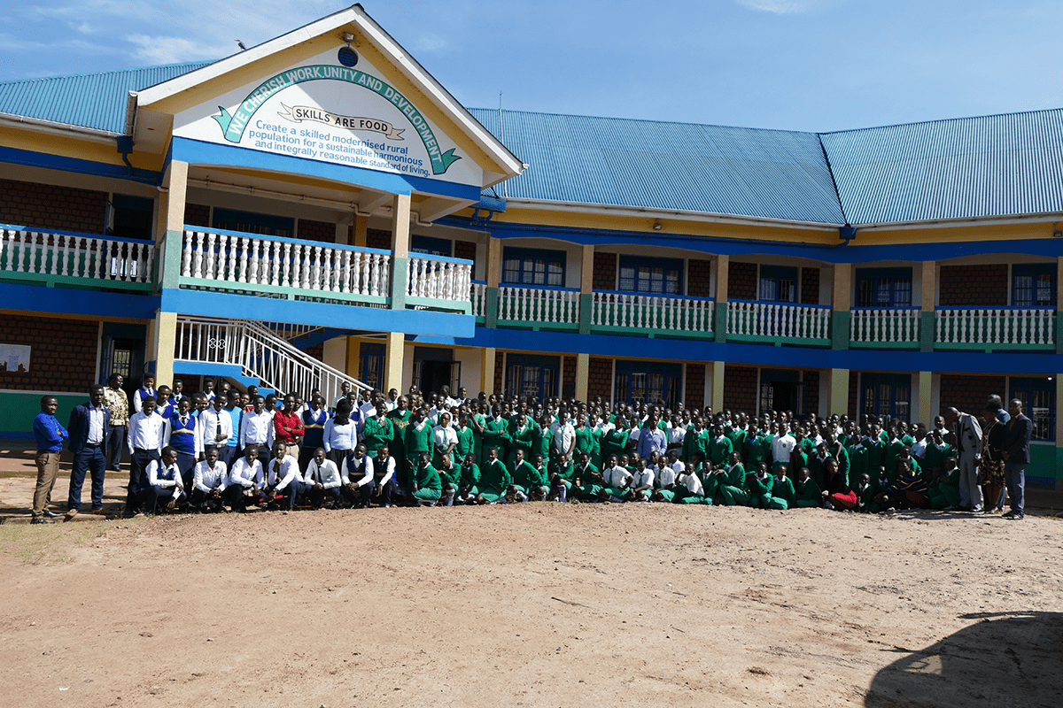 Großes Gruppenfoto von Schülern und Lehrern in grünen Uniformen vor dem Schulgebäude der St. Konrad Schule Uganda, das Motto „Skills are Food“ über dem Eingang.