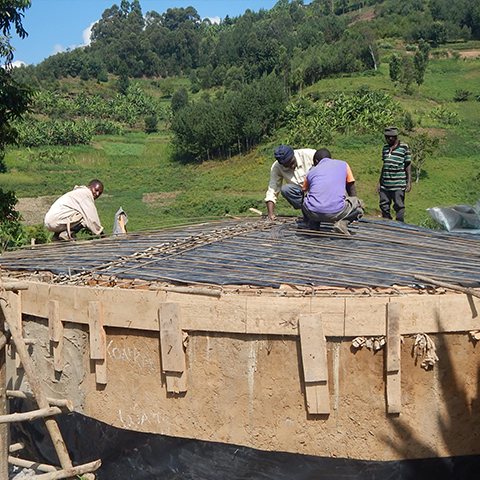 Arbeiter in Uganda beim Bau eines Wasserspeichers an der St. Konrad Schule, mit Stahlverstärkung und Holzschalung, umgeben von grüner Landschaft im Hintergrund.