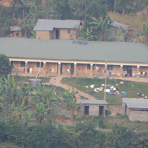 Schlafgebäude an der St. Konrad Schule in Uganda, mit aufgehängter Wäsche und umgeben von Pflanzen, Bäumen und kleineren Nebengebäuden in ländlicher Umgebung.