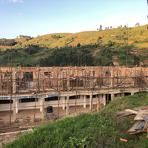 Baufortschritt des Irene Magdalena Santa Maria Workshops an der St. Konrad Schule Uganda, mit Gerüsten und hölzernen Baugerüsten, umgeben von grüner Landschaft.