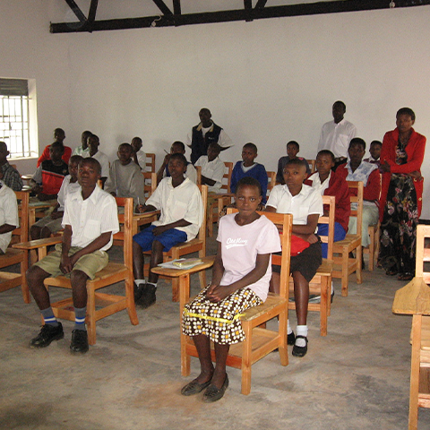 Schüler der St. Konrad Schule in Uganda sitzen in einem Klassenzimmer auf Holzstühlen und hören aufmerksam dem Lehrer zu, der vorne steht.