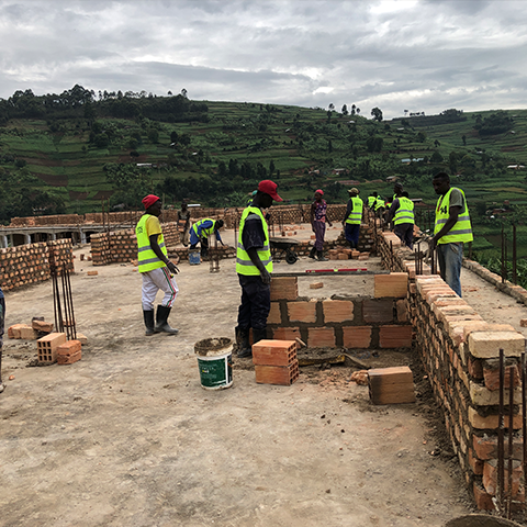 Arbeiter mit Warnwesten errichten Wände aus Ziegelsteinen im Rahmen eines Bauprojekts an der St. Konrad Schule in Uganda, mit grüner Landschaft im Hintergrund.