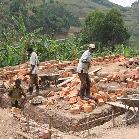 Arbeiter errichten ein neues Gebäude an der St. Konrad Schule Uganda, mit Ziegelsteinen, Schubkarren und Baumaterialien auf der Baustelle, umgeben von grüner Landschaft.