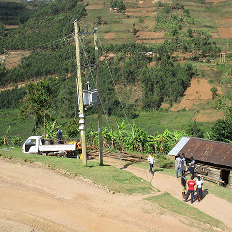 Techniker installieren Strommasten und Transformator an der St. Konrad Schule in Uganda, umgeben von ländlicher Landschaft; ein Truck und einige Schüler sind ebenfalls zu sehen.