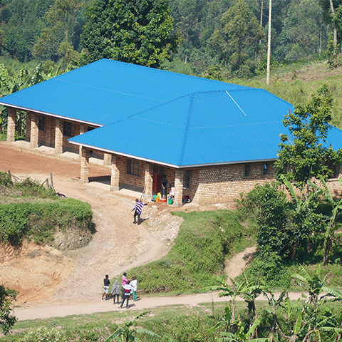 Neues Schulgebäude an der St. Konrad Schule Uganda mit leuchtend blauem Dach und umgeben von grüner Landschaft; Schüler und Lehrer vor dem Eingang.